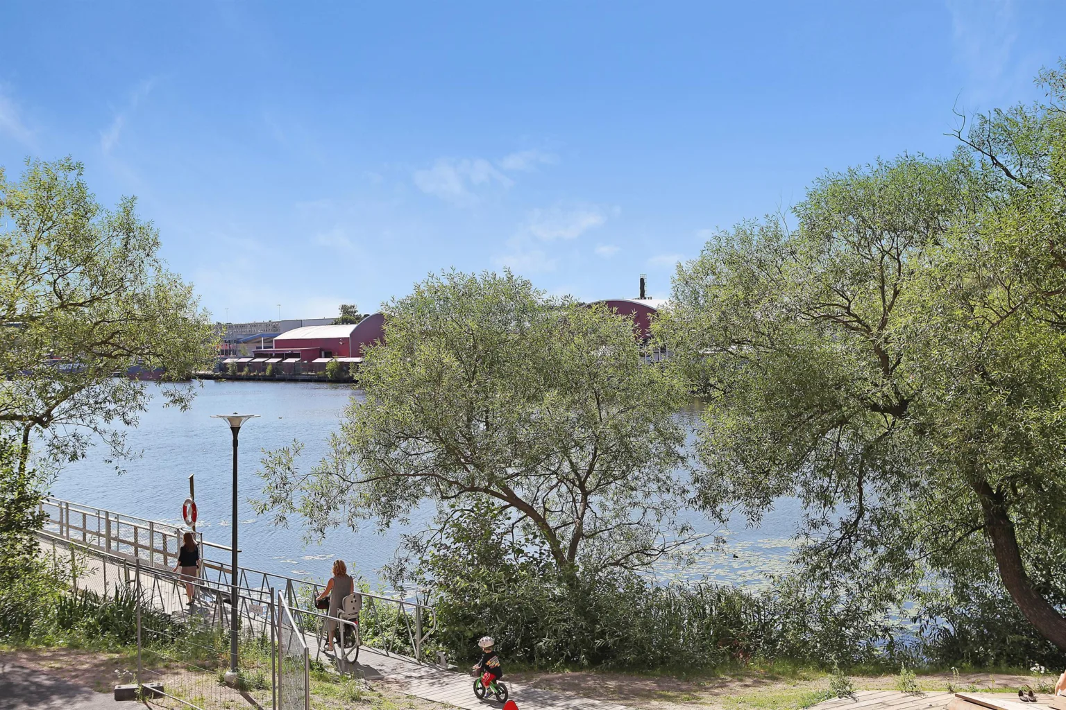 En flodkant med träd och buskar, en gångväg och människor som promenerar. I bakgrunden syns röda byggnader och blå himmel.