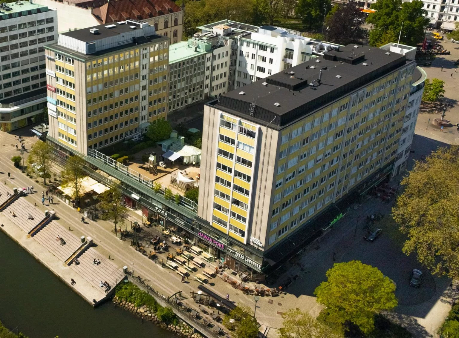 Flygbild av ett stort gult byggnadskomplex med uteserveringar vid vattnet.