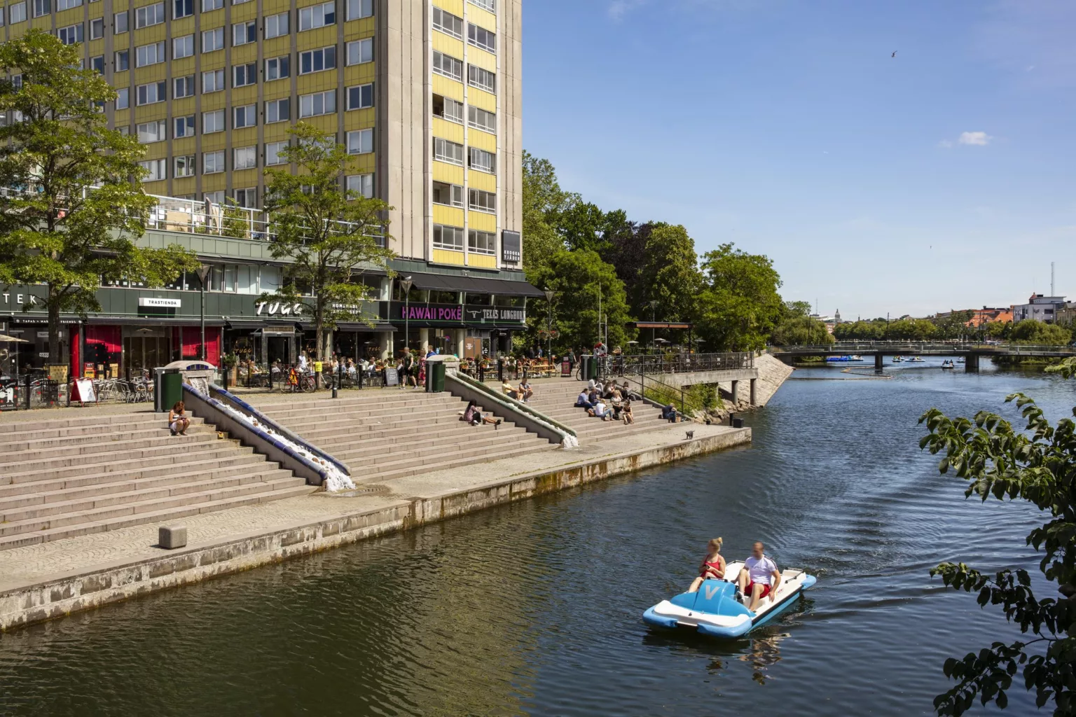 En trampbåt med två personer på en kanal nära en byggnad med trappor och uteserveringar.