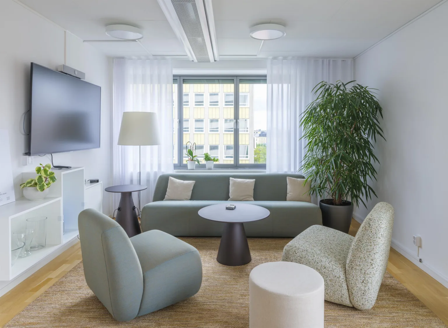 Ett vardagsrumsliknande kontorsutrymme med en soffa, fåtöljer, ett soffbord, en stor växt och gardiner vid fönstret.
