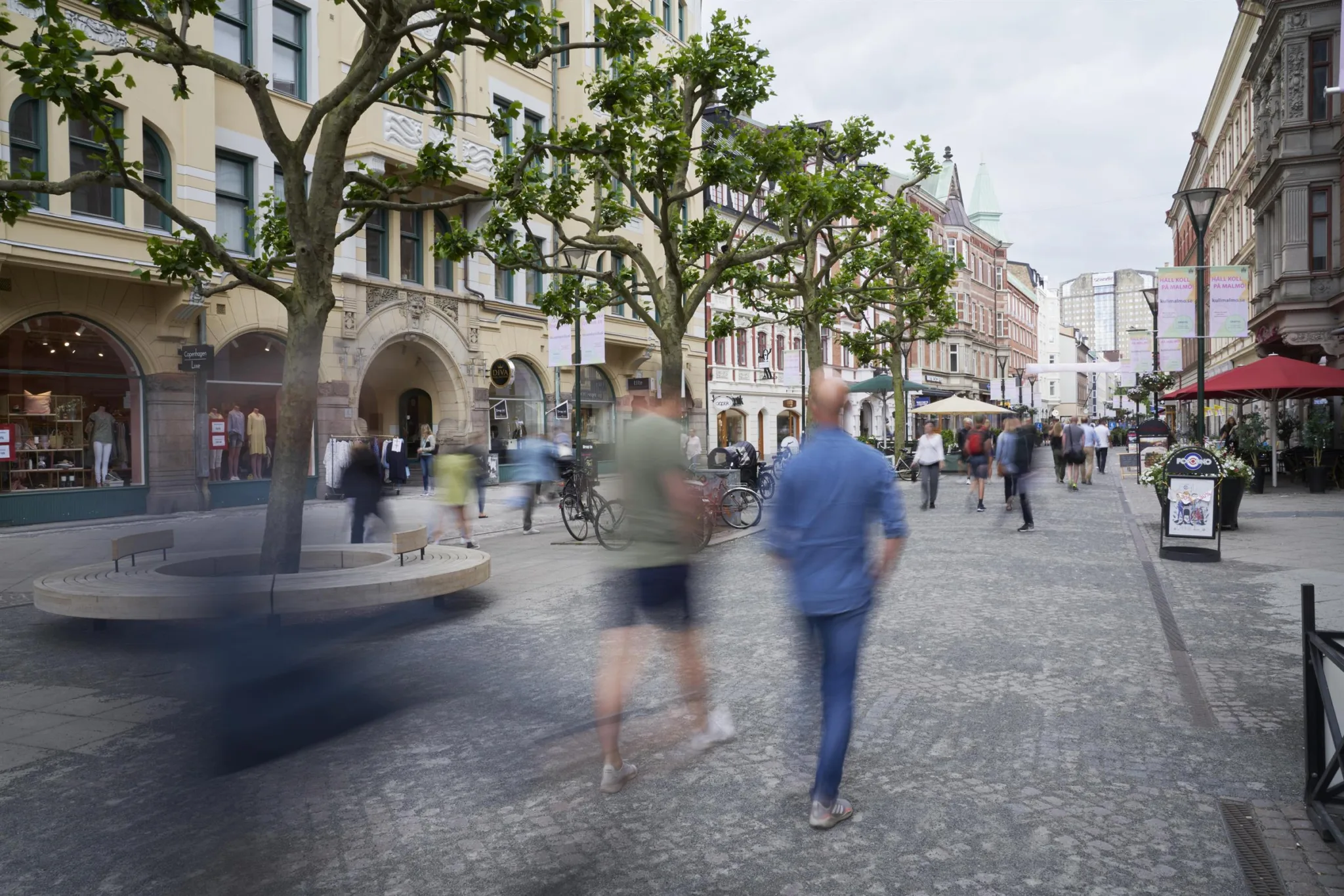 En stadsgata med människor som promenerar bland träd och byggnader.