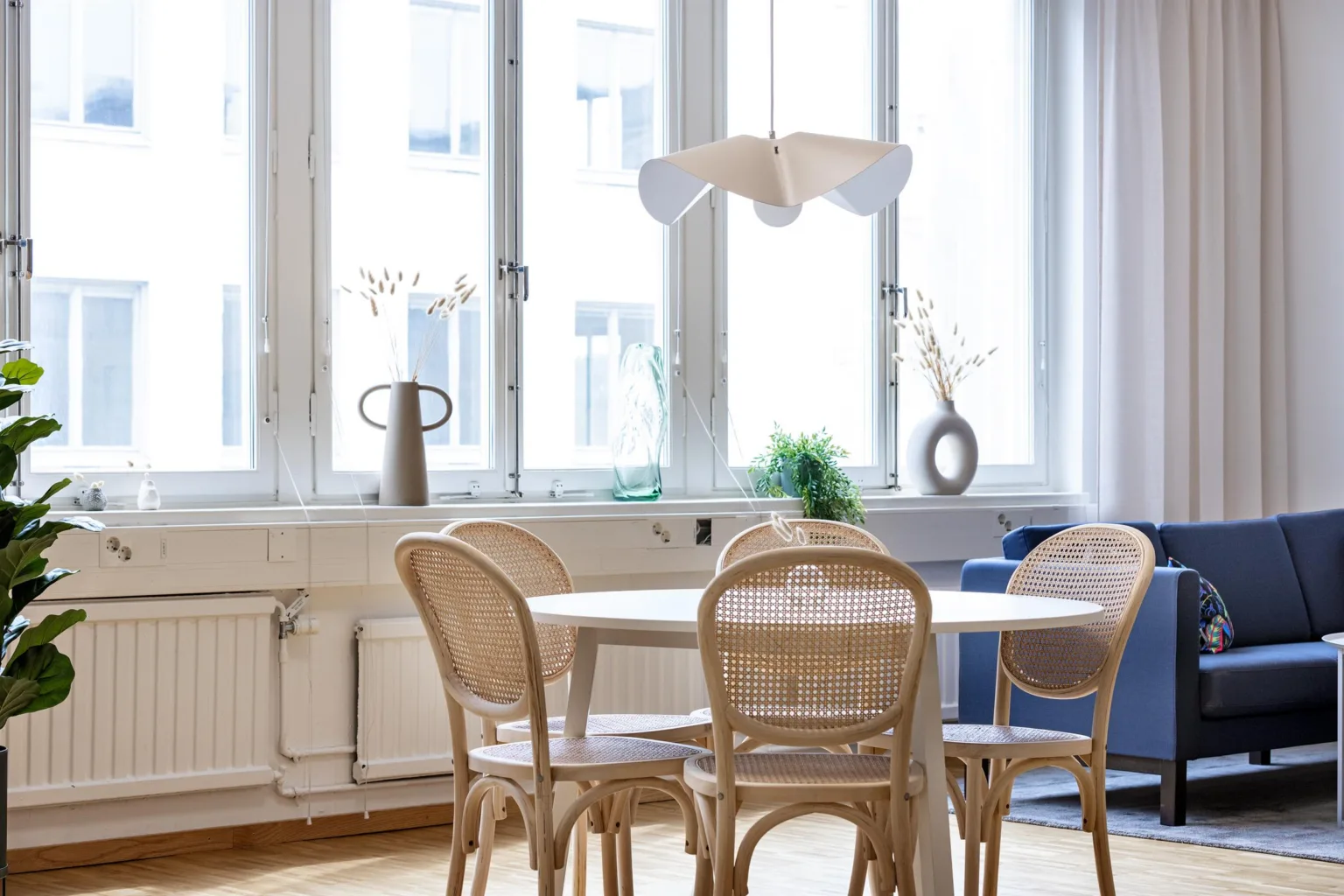 En matsal med trästolar och ett runt bord framför stora fönster, dekorerad med en modern lampa och små växter.