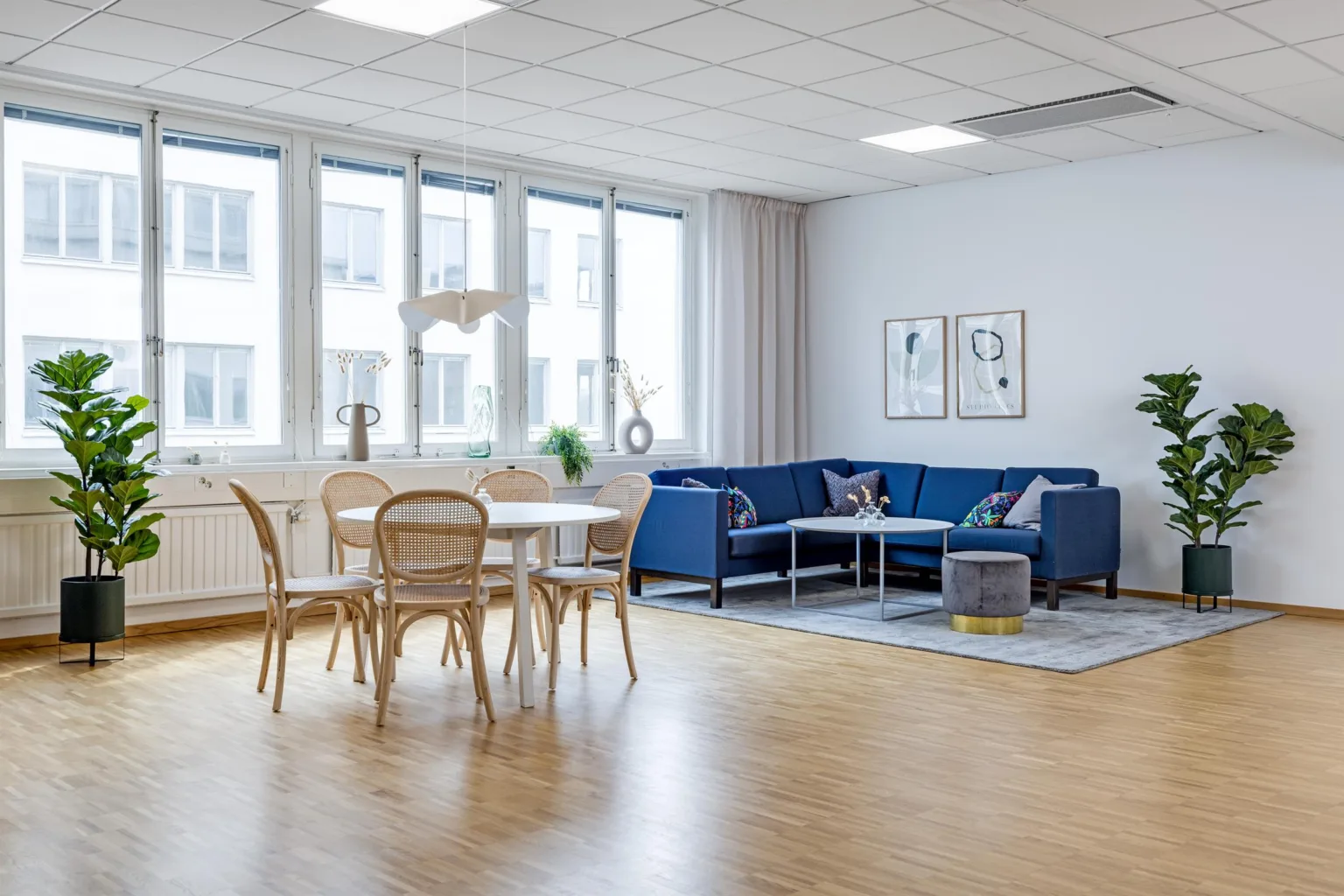 Ett ljust vardagsrum med blå soffa, vita matbord och rottingstolar på trägolv.