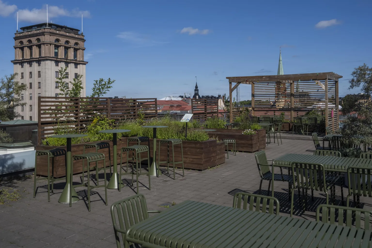 En takterrass med gröna bord och stolar, växtlådor samt utsikt över stadens byggnader och himmel.