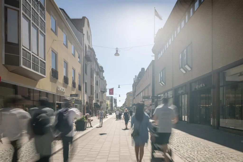 En solig gågata med människor som promenerar och byggnader på båda sidor.