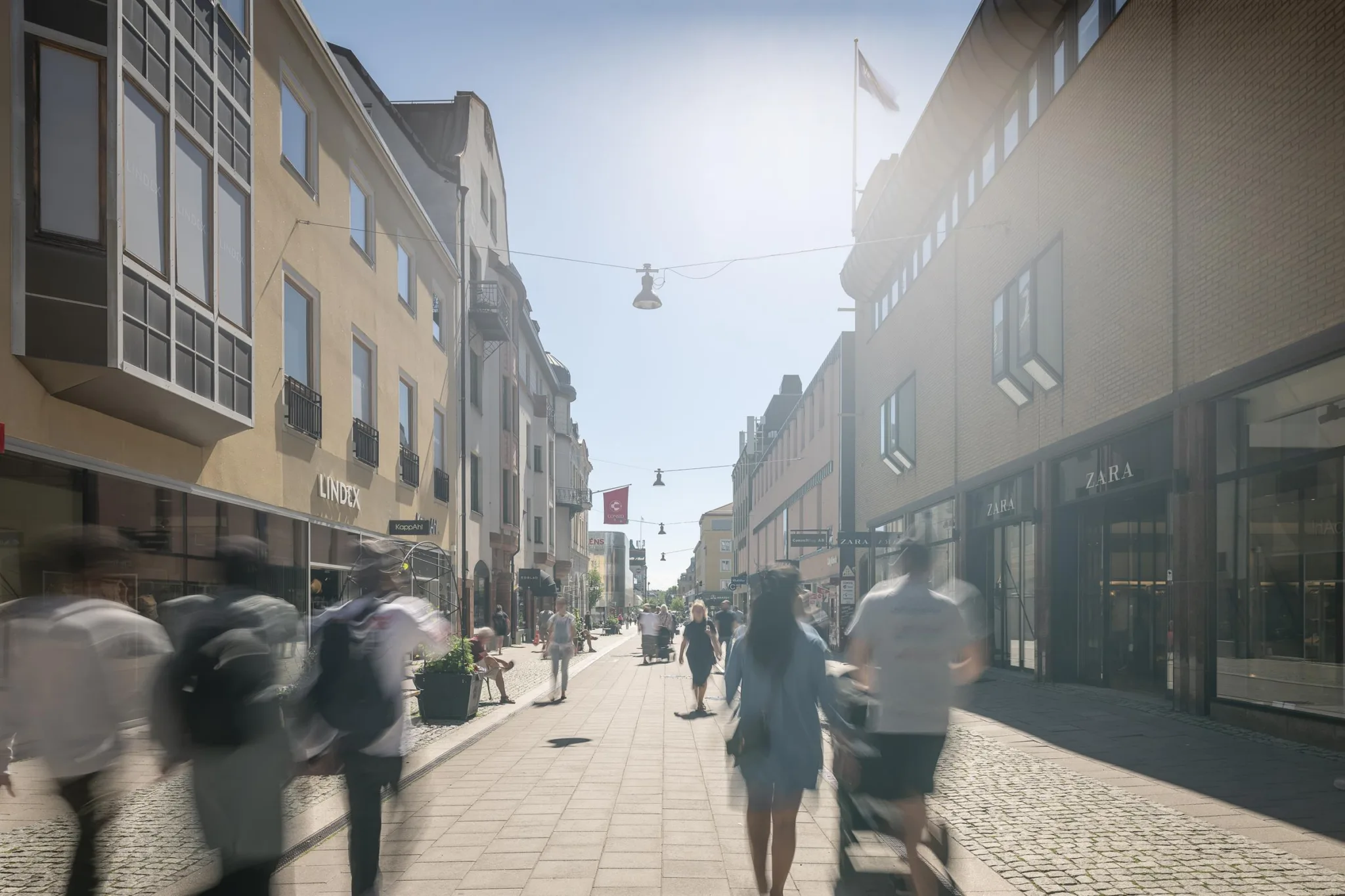 En solig gågata med människor som promenerar och byggnader på båda sidor.