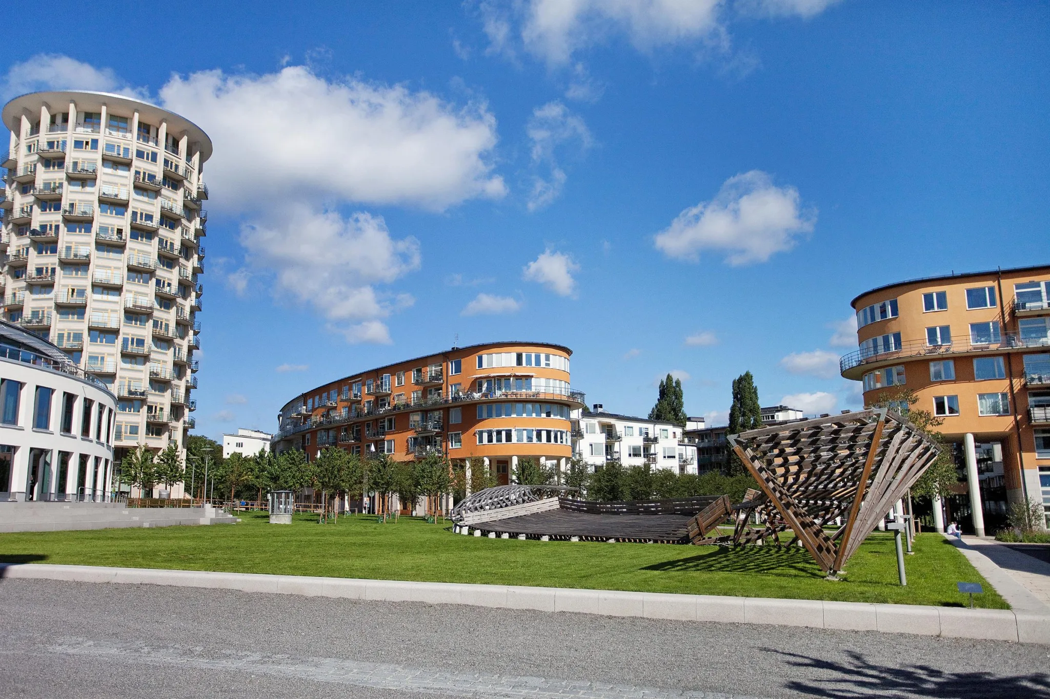 En utomhusmiljö med moderna flerfamiljshus, grön gräsmatta och en träkonstruktion.