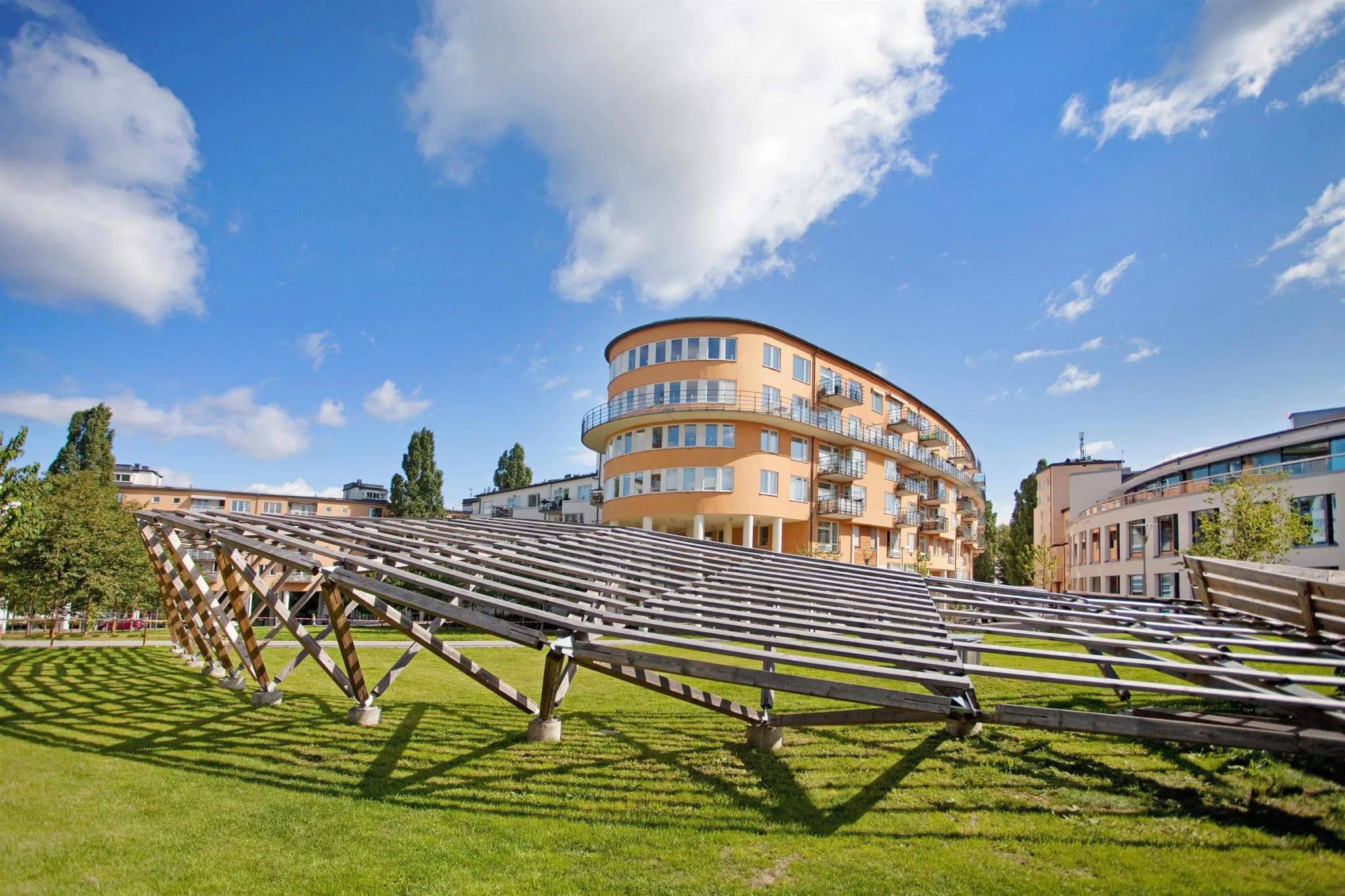 En solig park med en träkonstruktion i förgrunden och ett modernt bostadshus i bakgrunden.