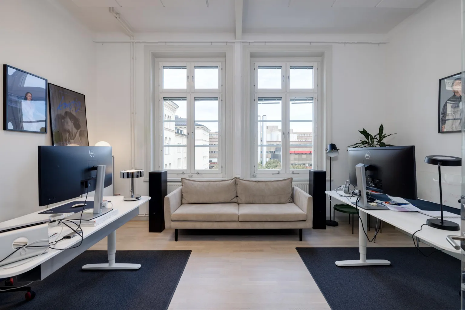 Ett kontorsutrymme med skrivbord och datorer på vardera sida, en beige soffa i mitten och stora fönster i bakgrunden.