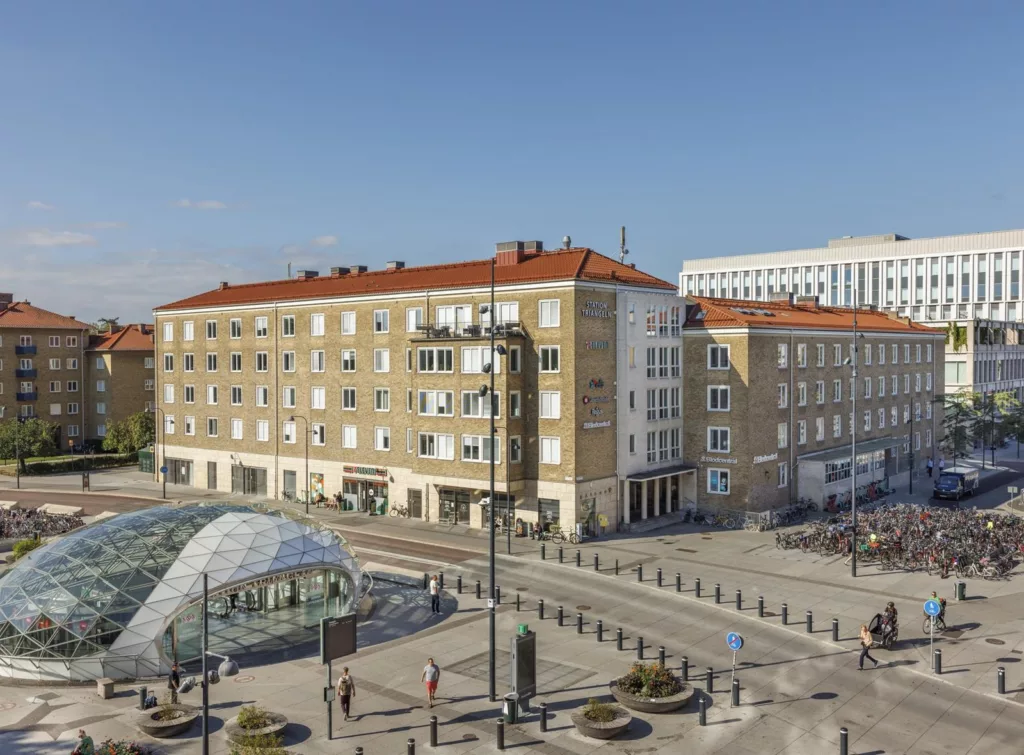 En öppen stadsplats med ett glaskupolformat tak, omgivna av byggnader med röda tak och blå himmel.