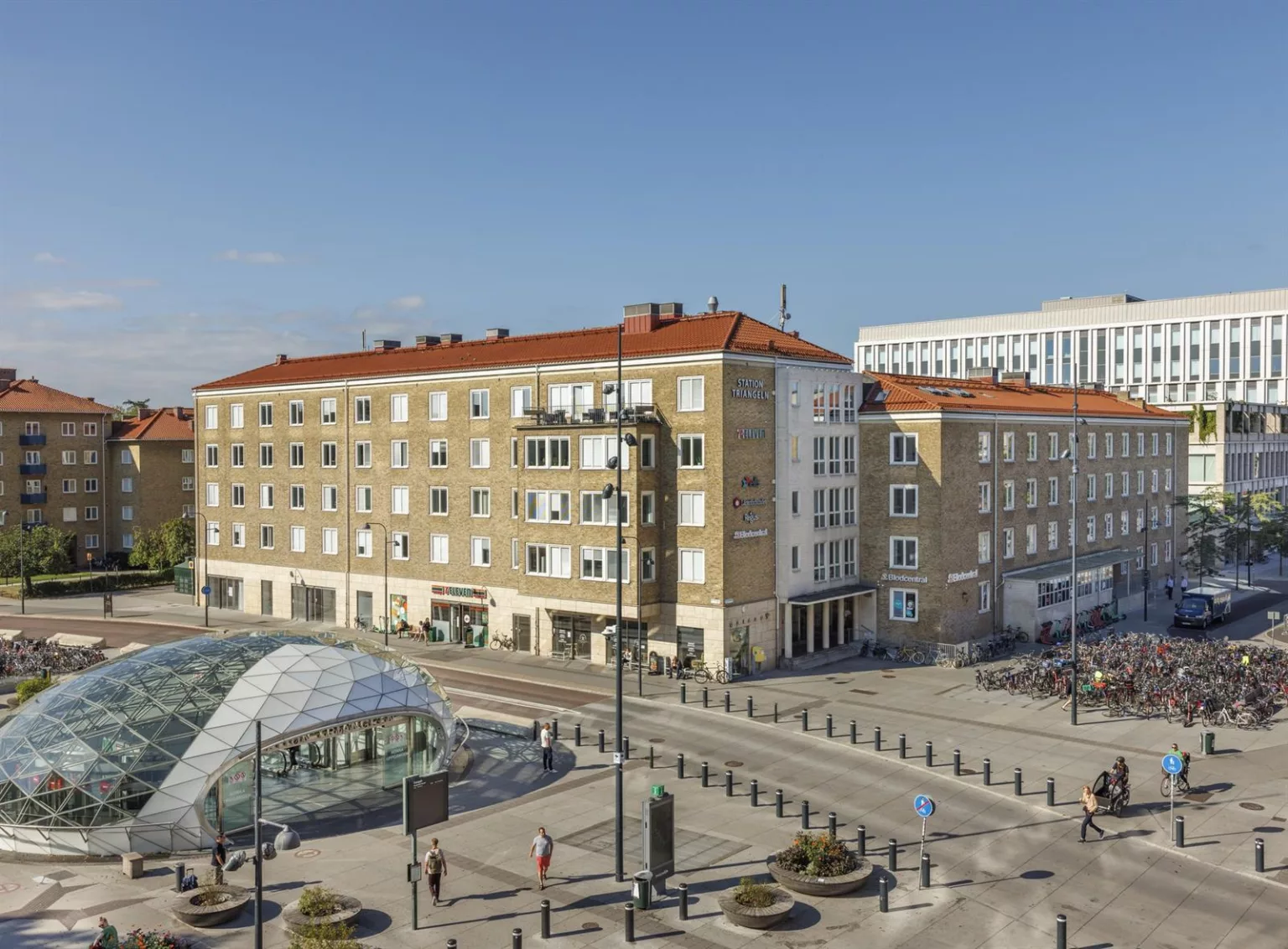 En öppen stadsplats med moderna byggnader, en glasstruktur och gångvägar med trafikskyltar och fotgängare.