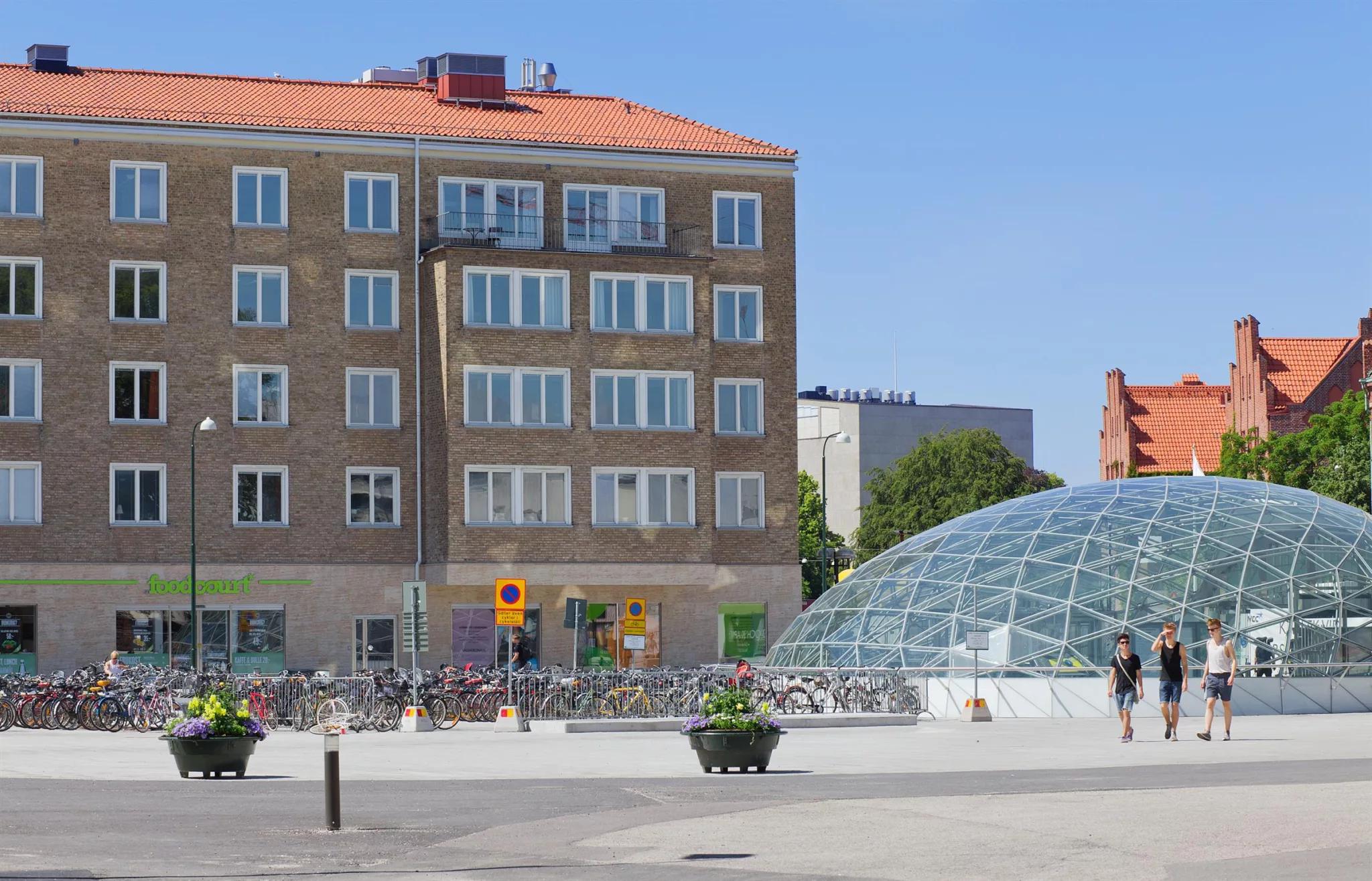 4. Glasbyggnad på ett torg nära ett flerbostadshus med tegelfasad, cykelställ och grönska i bakgrunden.