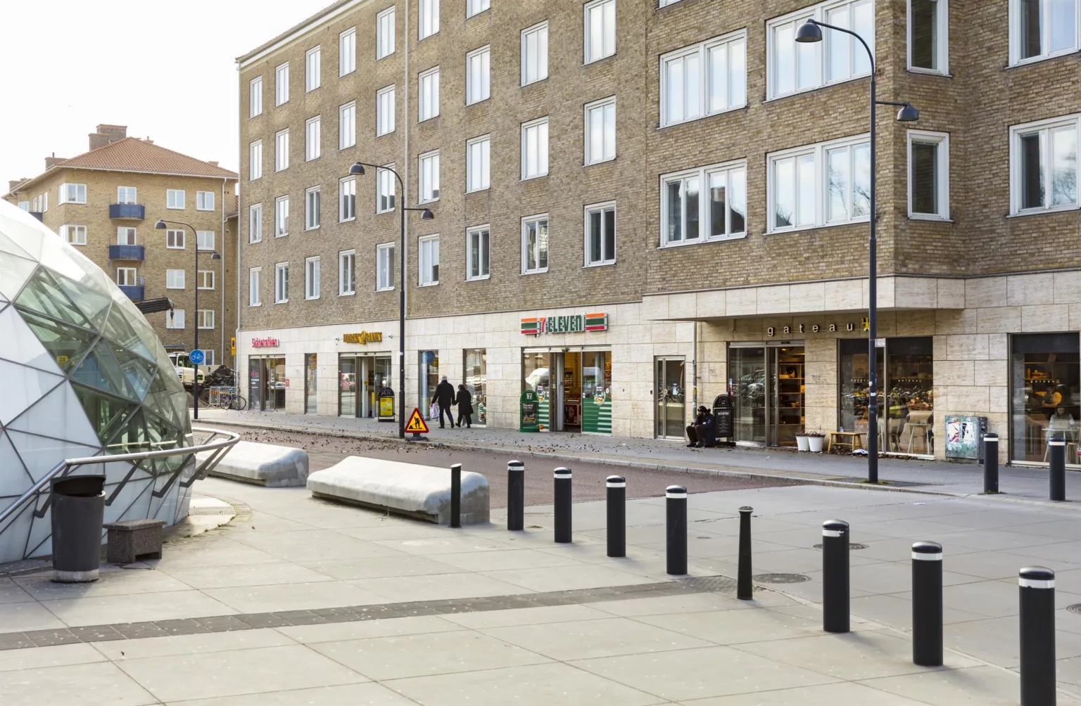 3. Flerbostadshus med butiker på bottenvåningen, bred trottoar med cykelställ och pollare.