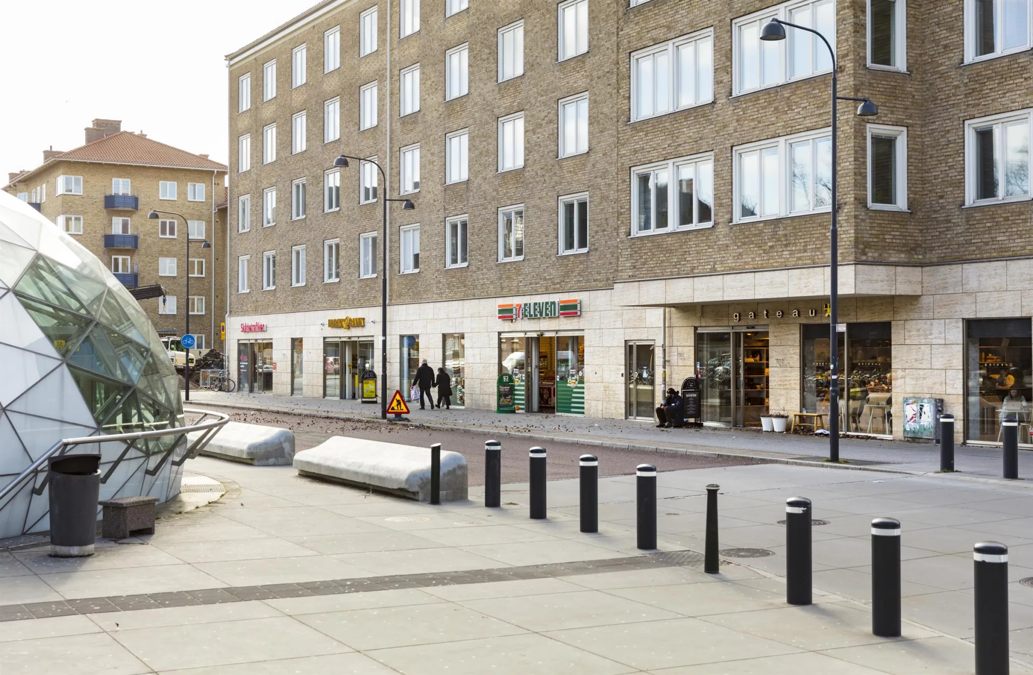 3. Flerbostadshus med butiker på bottenvåningen, bred trottoar med cykelställ och pollare.