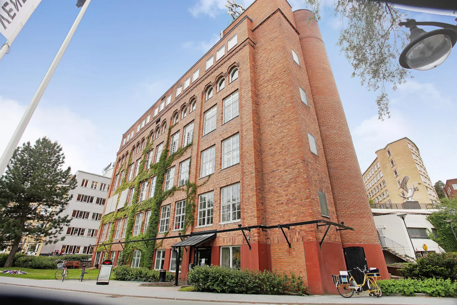 En hög röd tegelbyggnad med gröna klängväxter, omgiven av mindre byggnader och träd.