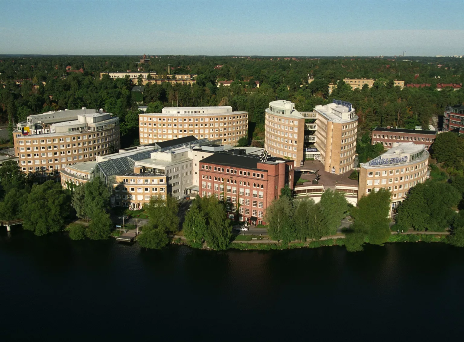 Flera byggnader i olika storlekar vid en vattenkant, omgivna av träd och grönska.