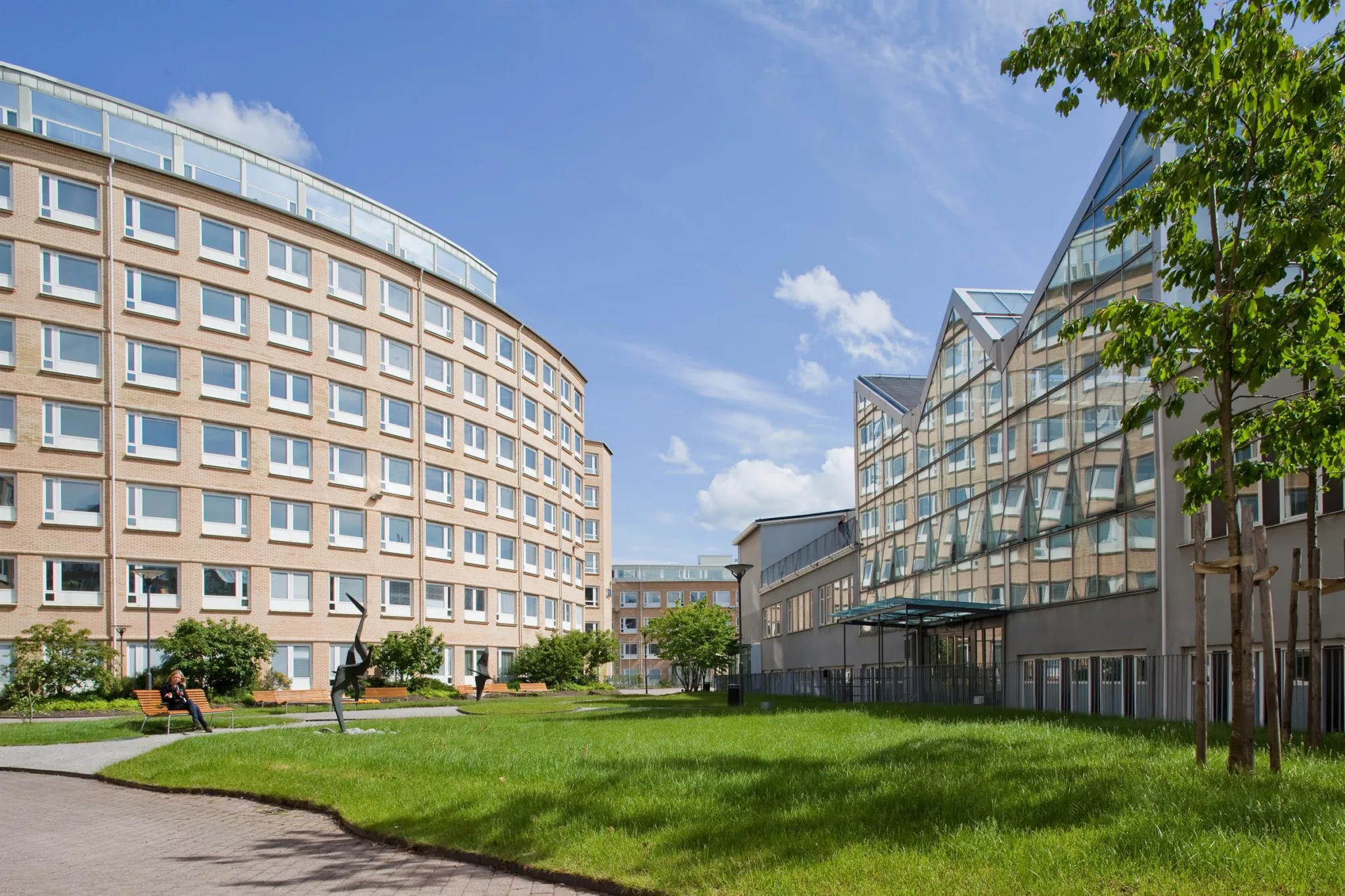 Två stora byggnader med modern arkitektur, omgivna av grön gräsmatta och träd.