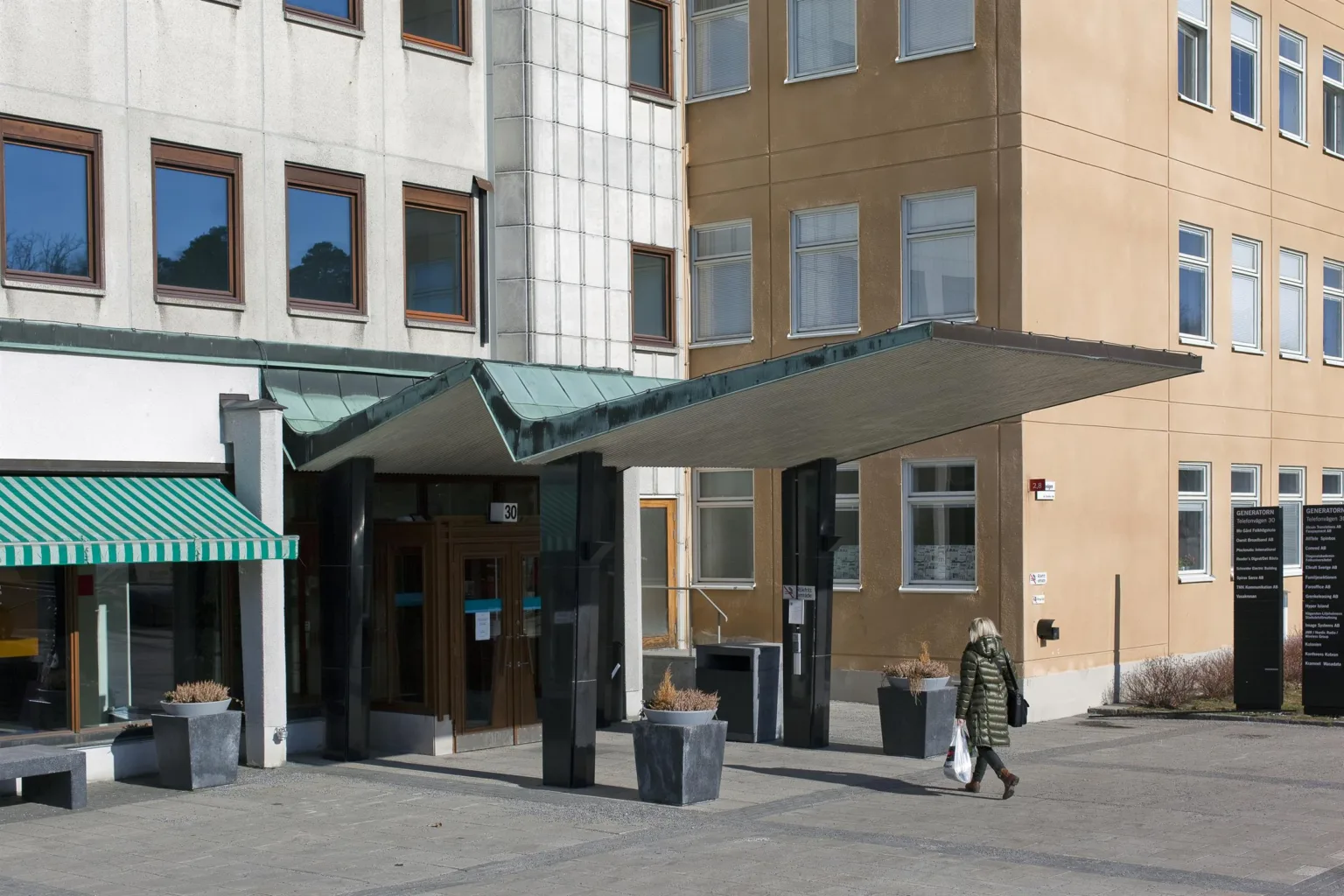 Entré till en byggnad med en grön baldakin, omgiven av moderna fasader och en gående person.