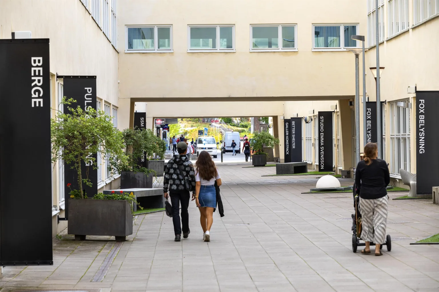 Personer promenerar genom en gång med planteringar och moderna byggnader.