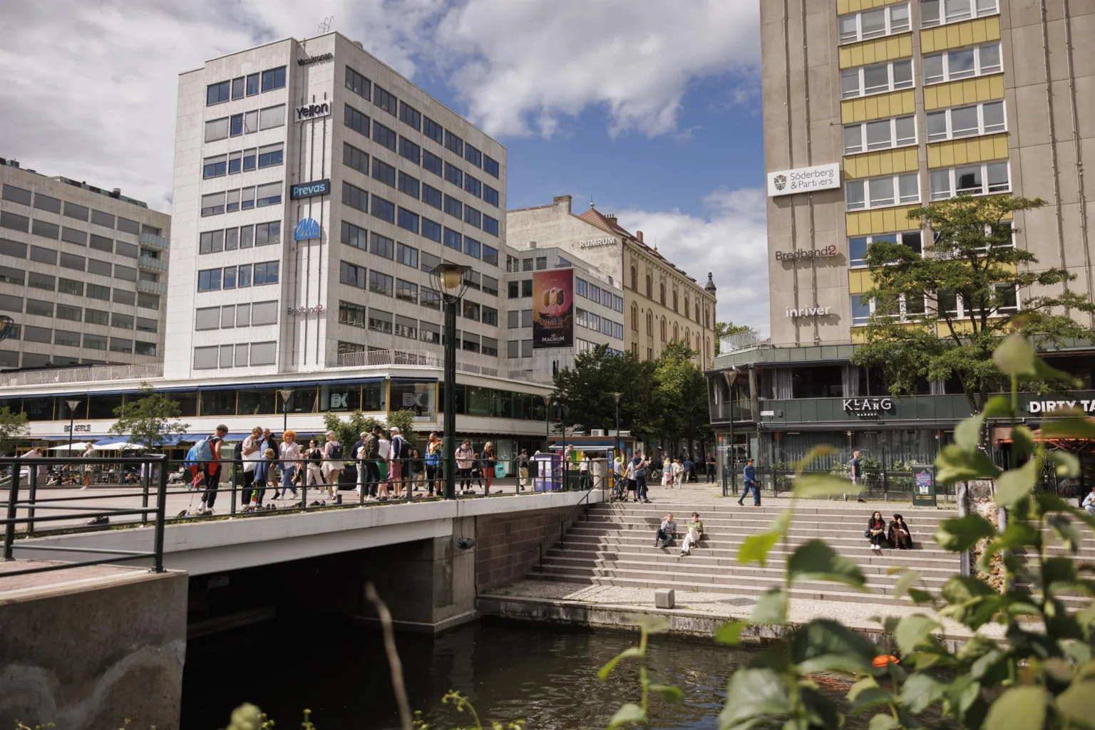 En stadsvy med människor vid en kanal och moderna byggnader omgivna av grönska under en klar himmel.