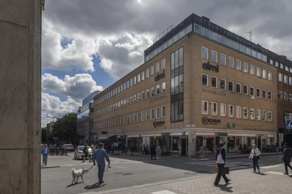 En hörnbyggnad med beige fasad, affärer på bottenvåningen och människor som går på gatan under molnig himmel.