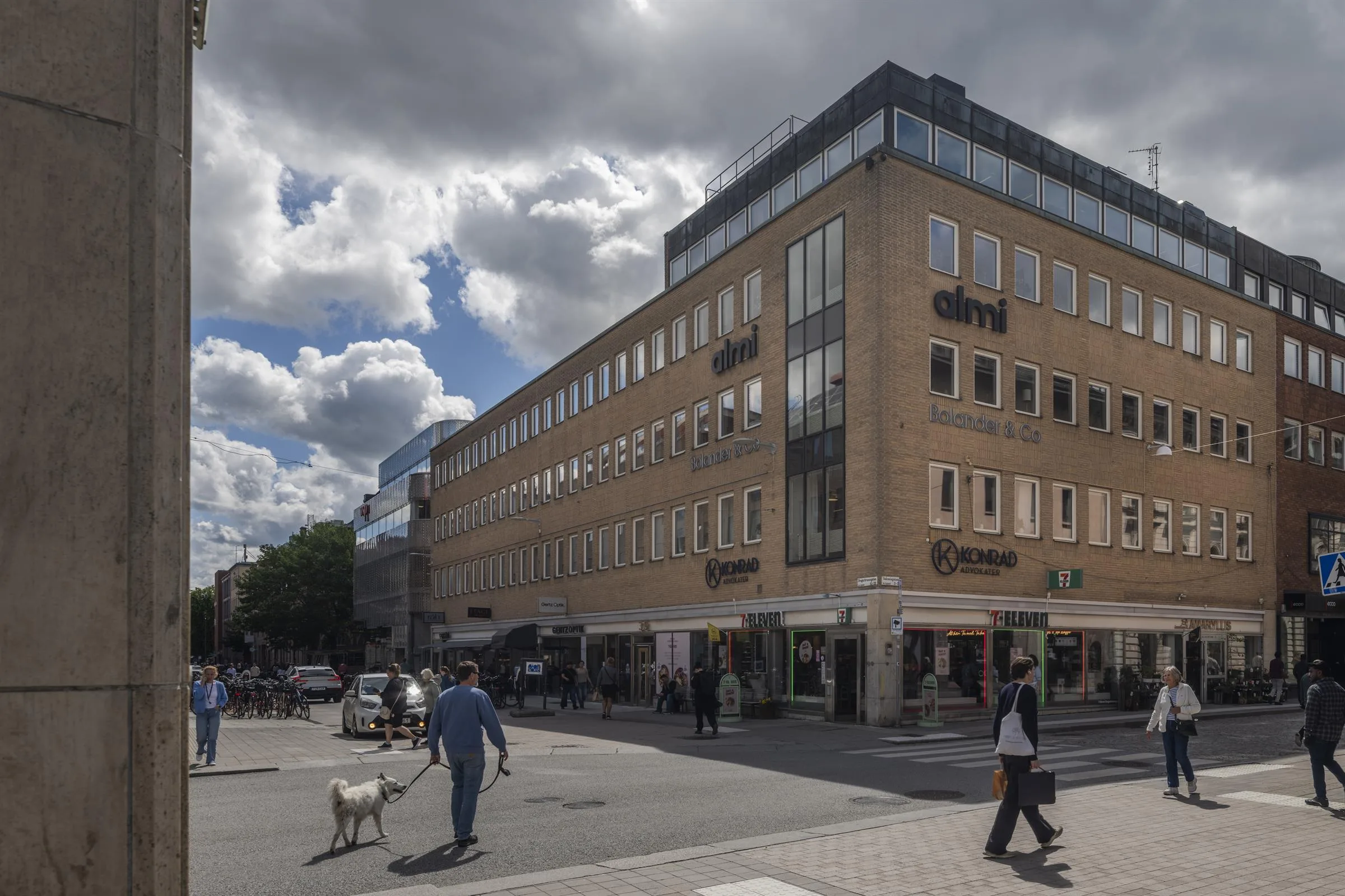 En hörnbyggnad med beige fasad, affärer på bottenvåningen och människor som går på gatan under molnig himmel.