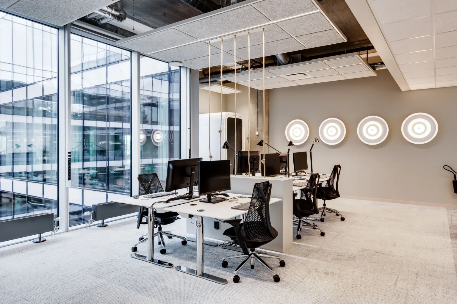 Ett modernt kontor med skrivbord, kontorsstolar och datorskärmar, omgiven av glasväggar med utsikt över en byggnad utanför.