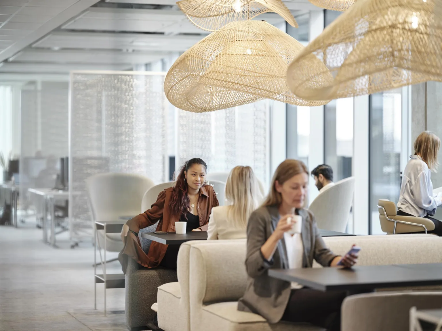 En kontorslounge med personer som sitter i soffor och vid bord under dekorativa taklampor.