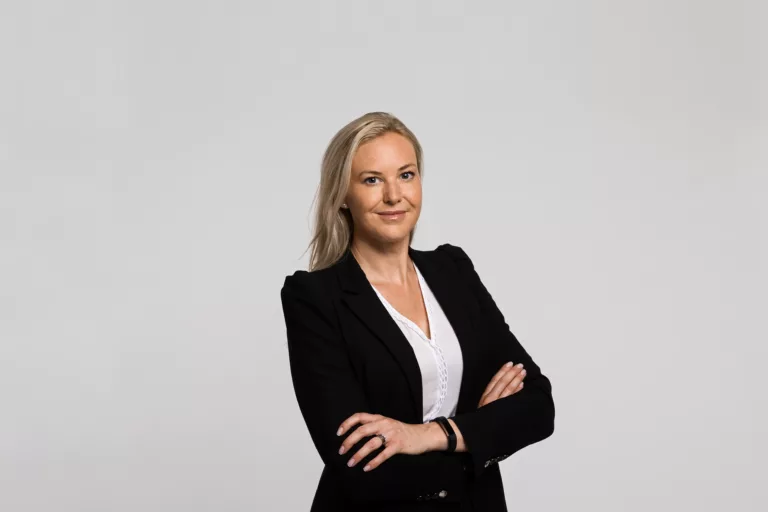 3. Kvinna i svart kavaj och vit blus står med armarna i kors mot en ljusgrå bakgrund.