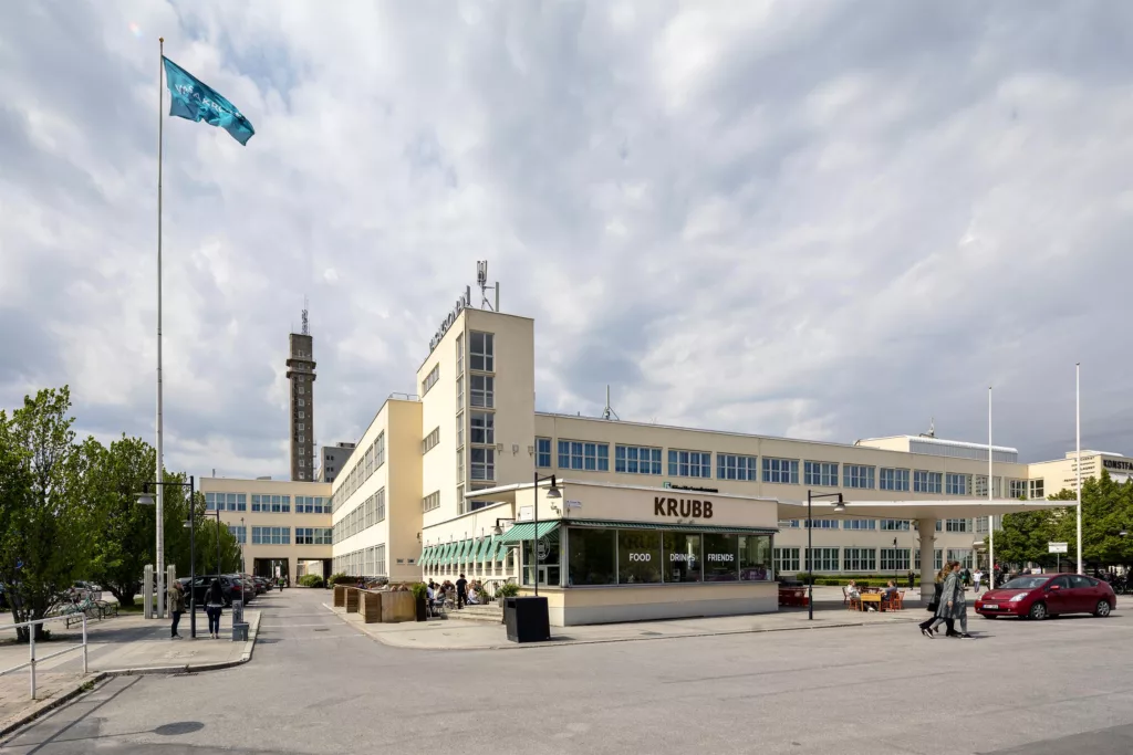 En byggnad med modern arkitektur, en flagga och ett högt torn syns i bakgrunden under en molnig himmel.