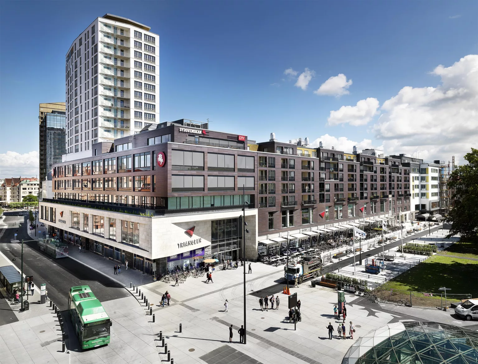 Ett stort torg med höga byggnader, en grön spårvagn och flera fotgängare.