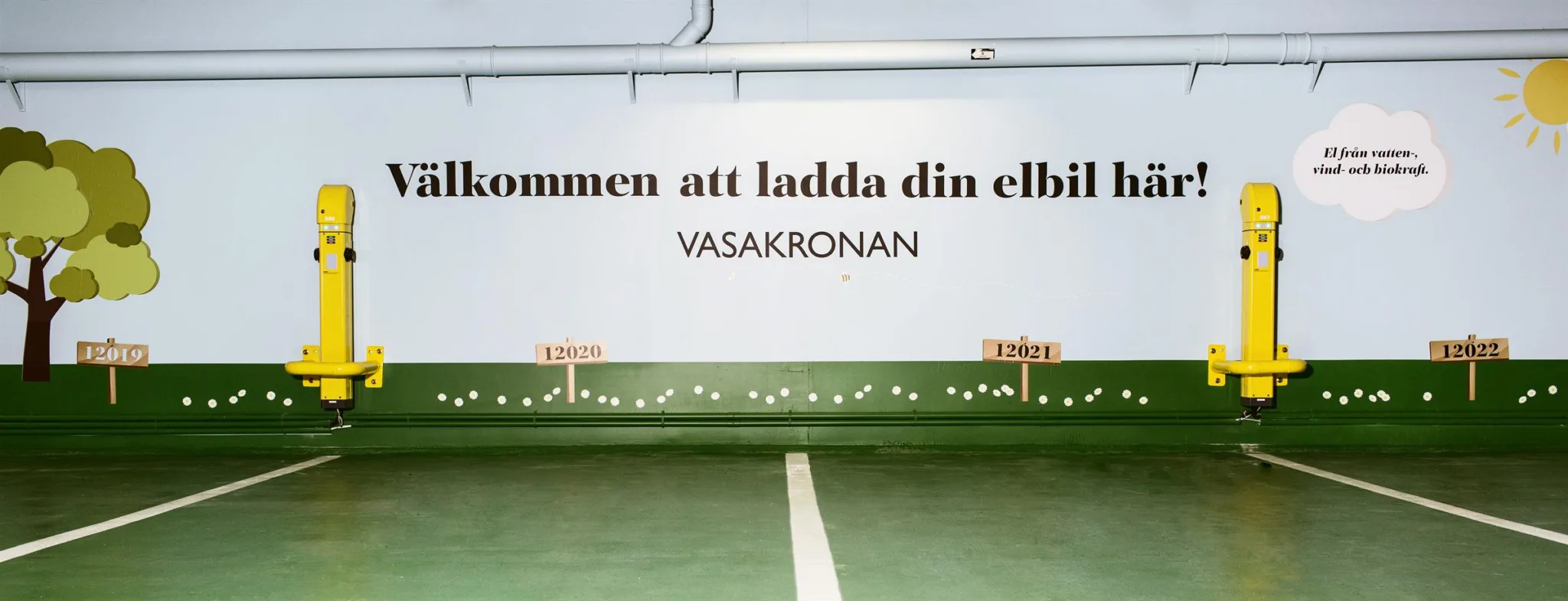 1. Vit vägg med texten "Välkommen att ladda din elbil" och "Vasakronan" ovanför en grön parkeringsyta.