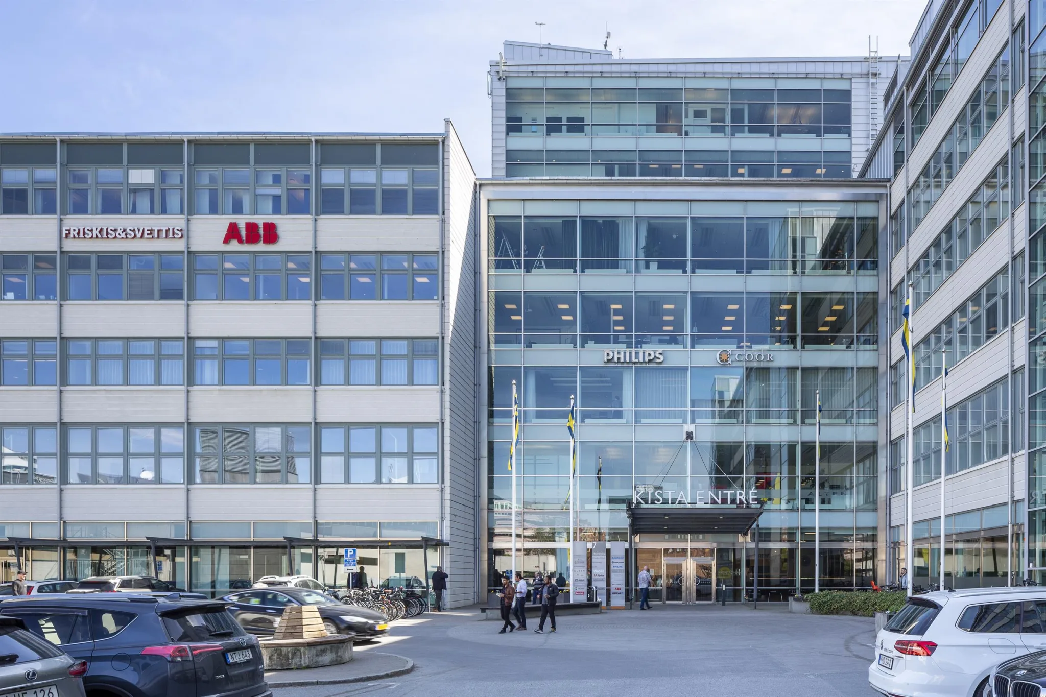 4. En byggnad med glasfasad och en ABB-logotyp på en angränsande byggnad, bilar och människor vid entrén.