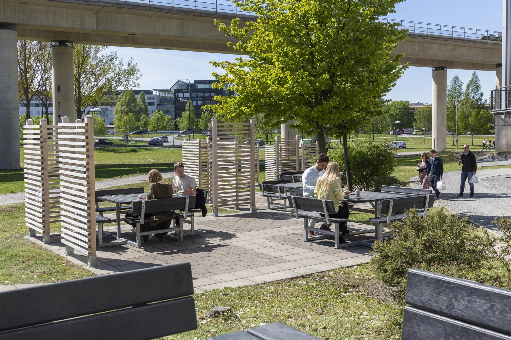 5. Ett utomhusområde med parkbänkar och bord, där människor sitter under träd och nära en gångbro.