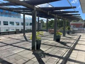 En takterrass med stenplattor, pergola och blomkrukor, omgiven av byggnader och grönska under en klar blå himmel.