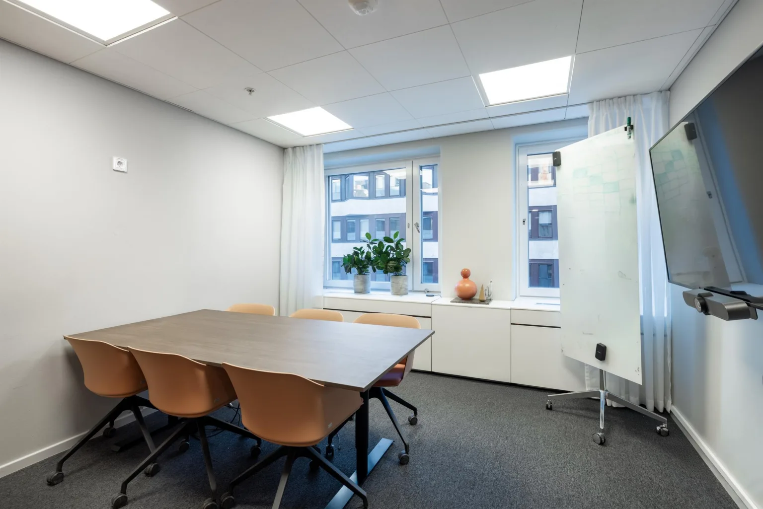 Ett mötesrum med ett stort bord, flera stolar, en whiteboard och fönster med utsikt mot en byggnad.