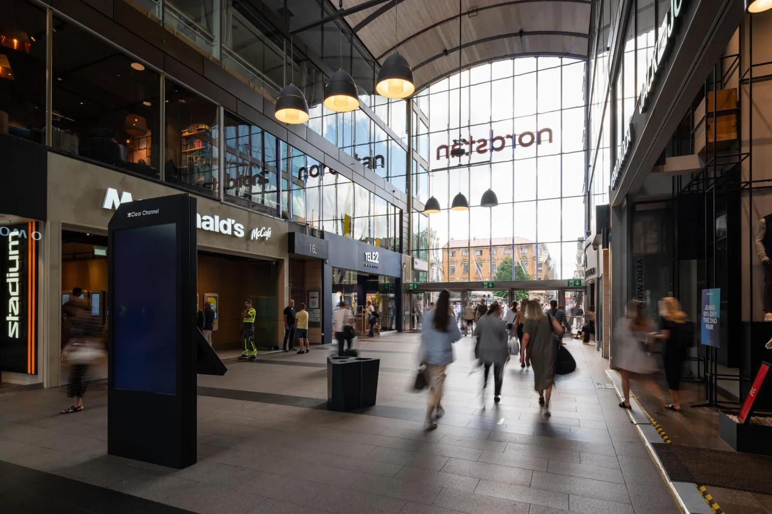 Ingång till en galleria med butiker, skyltar och människor som promenerar under glastak.