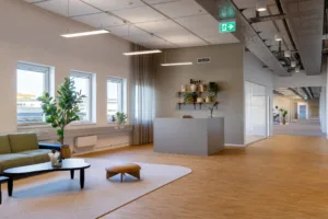 En reception med ett diskret skrivbord, växter och dekorativa hyllor, omgiven av stora fönster och trägolv.