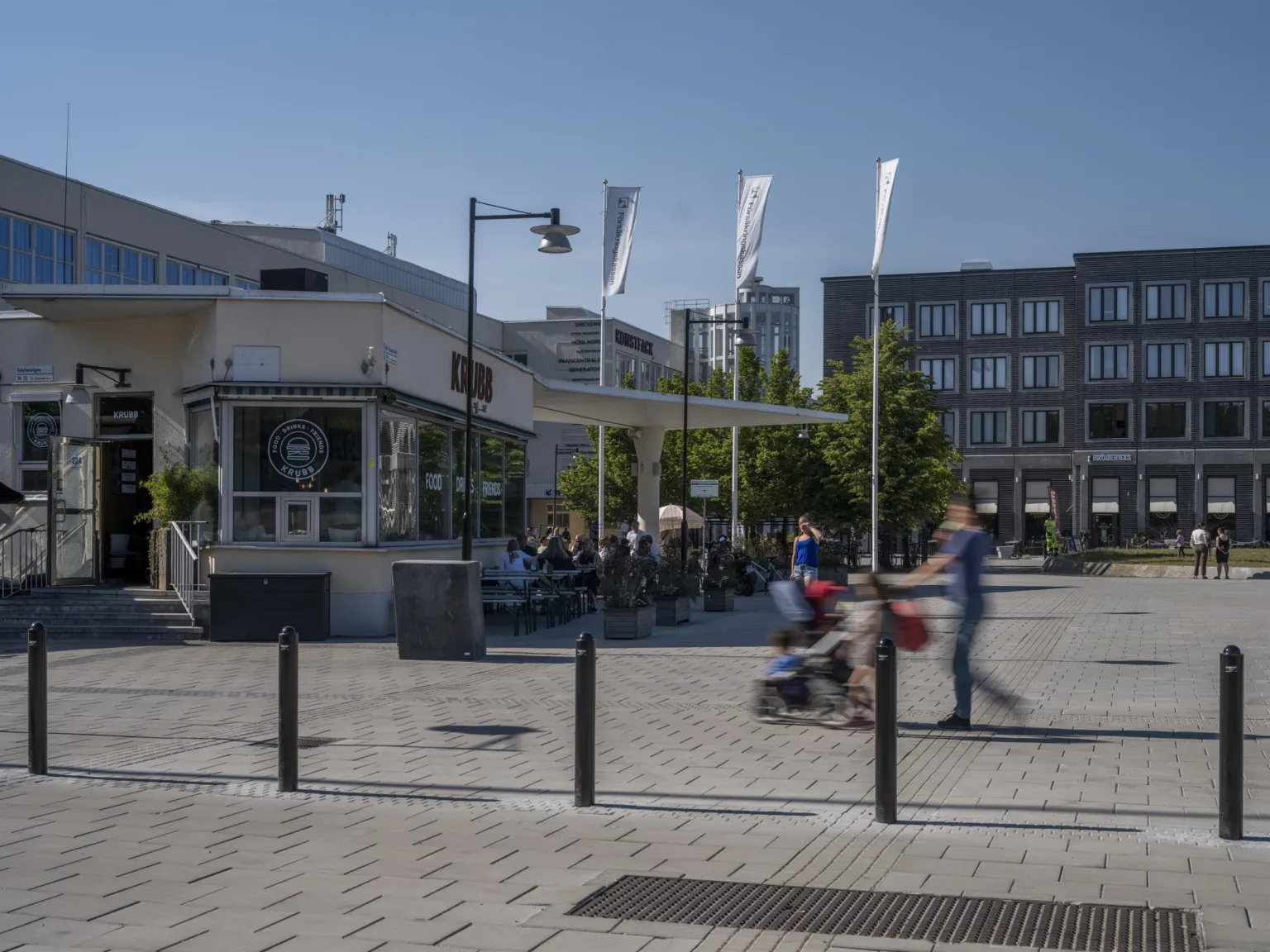5. Ett stadstorg med människor som går och en person som skjuter en barnvagn, omgiven av moderna byggnader, ett café och flaggor som vajar.