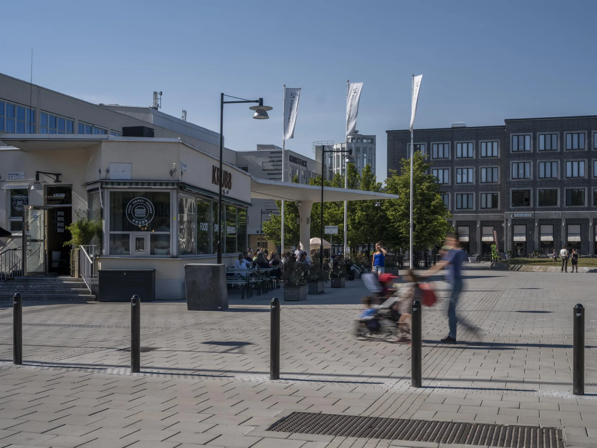 5. Ett stadstorg med människor som går och en person som skjuter en barnvagn, omgiven av moderna byggnader, ett café och flaggor som vajar.