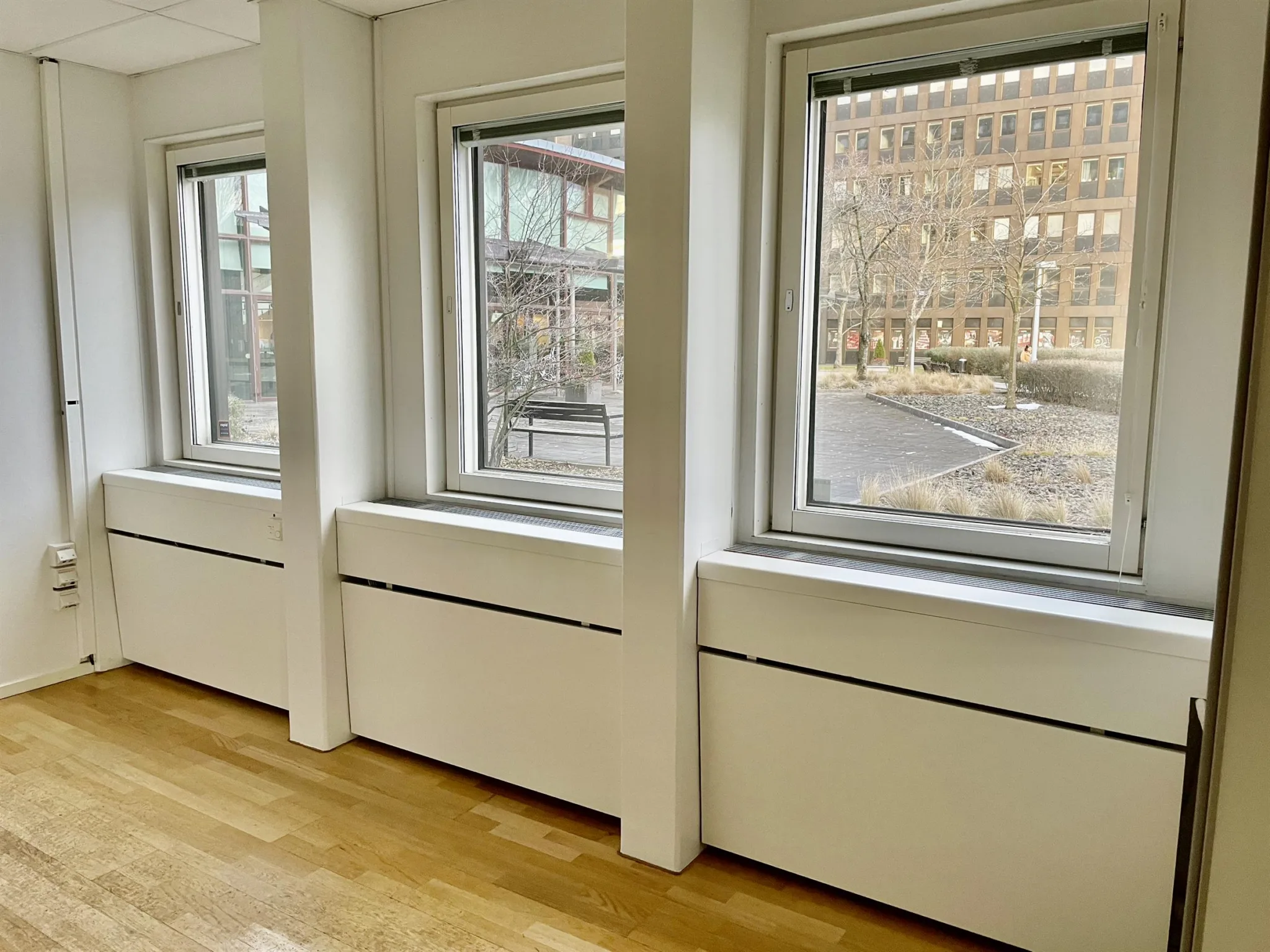 Interiör med stora fönster som vetter mot en innergård, trägolv och vita väggar.