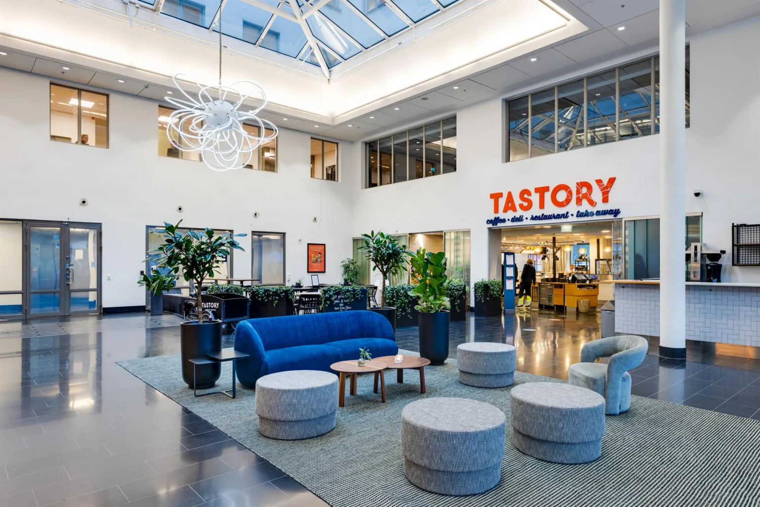 Ett rymligt atrium med sittmöbler, växter och en skylt för "Tastory" vid en restaurangentré.