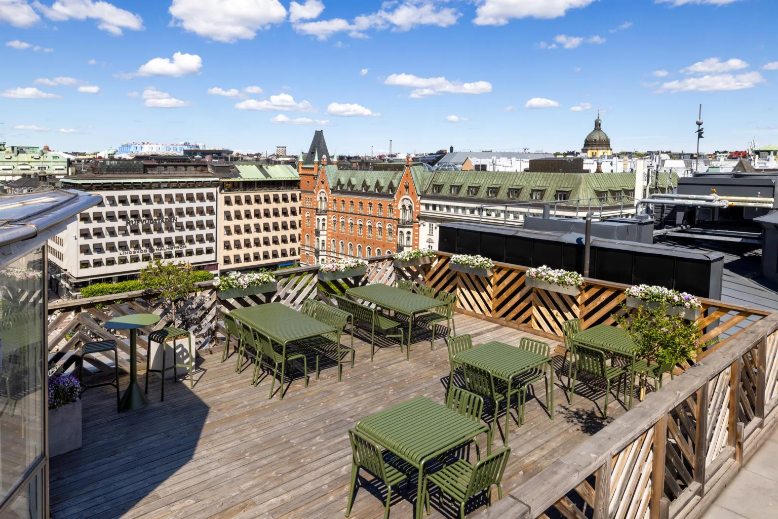 Takterrass med gröna bord och stolar, omgiven av byggnader under en klarblå himmel.