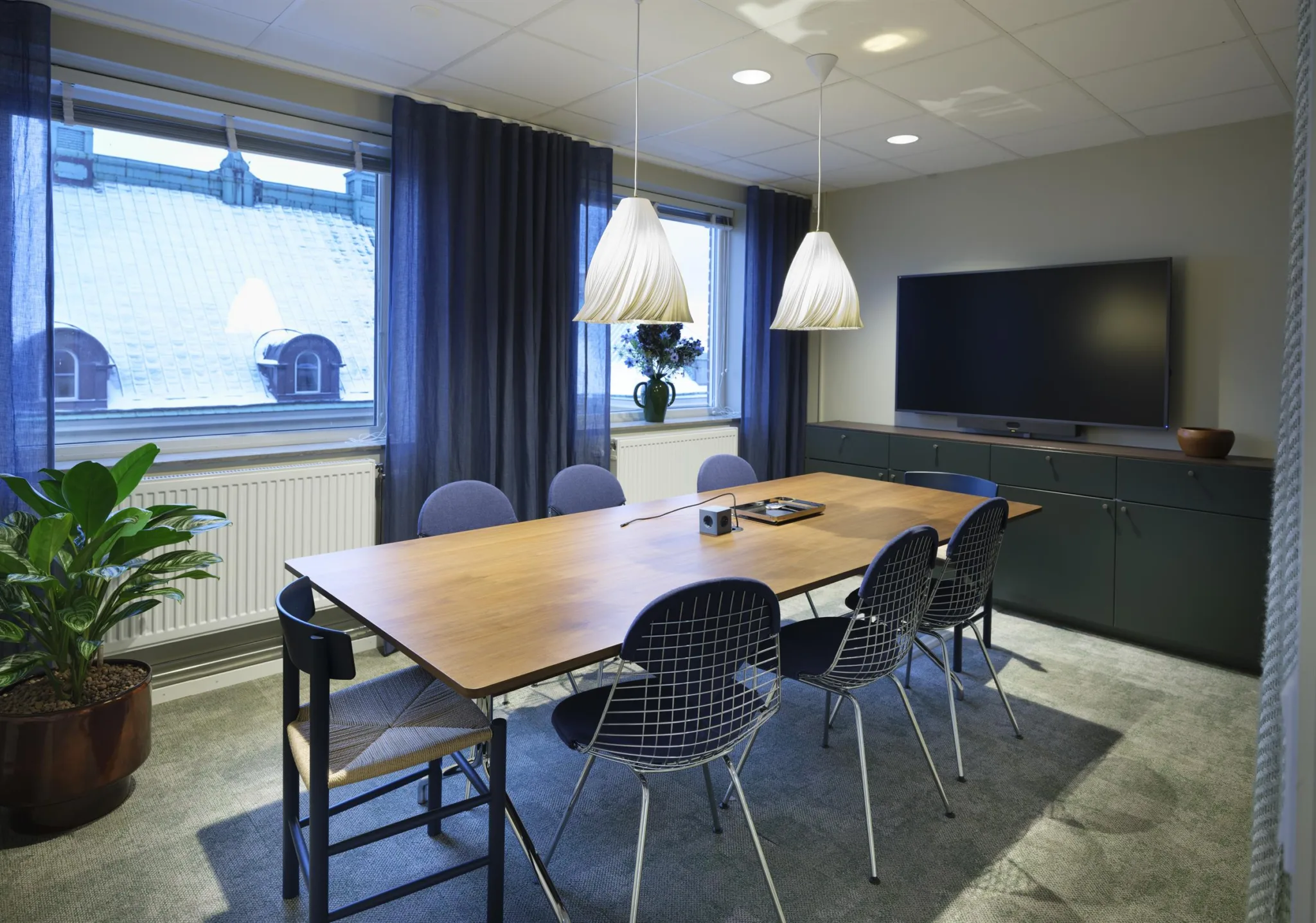 Ett mötesrum med ett långt träbord, sex stolar, två hängande lampor, en stor TV och fönster med mörka gardiner.