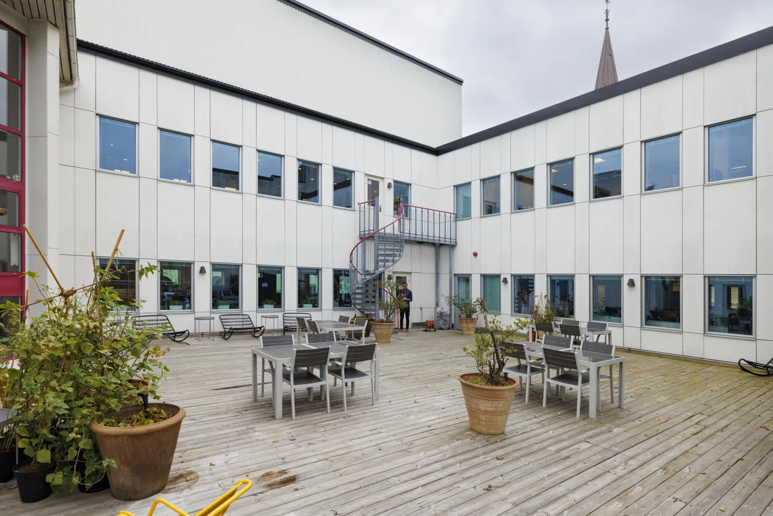 3. En utomhusgård med trämöbler, stora krukväxter och omgiven av en vit byggnad med många fönster.