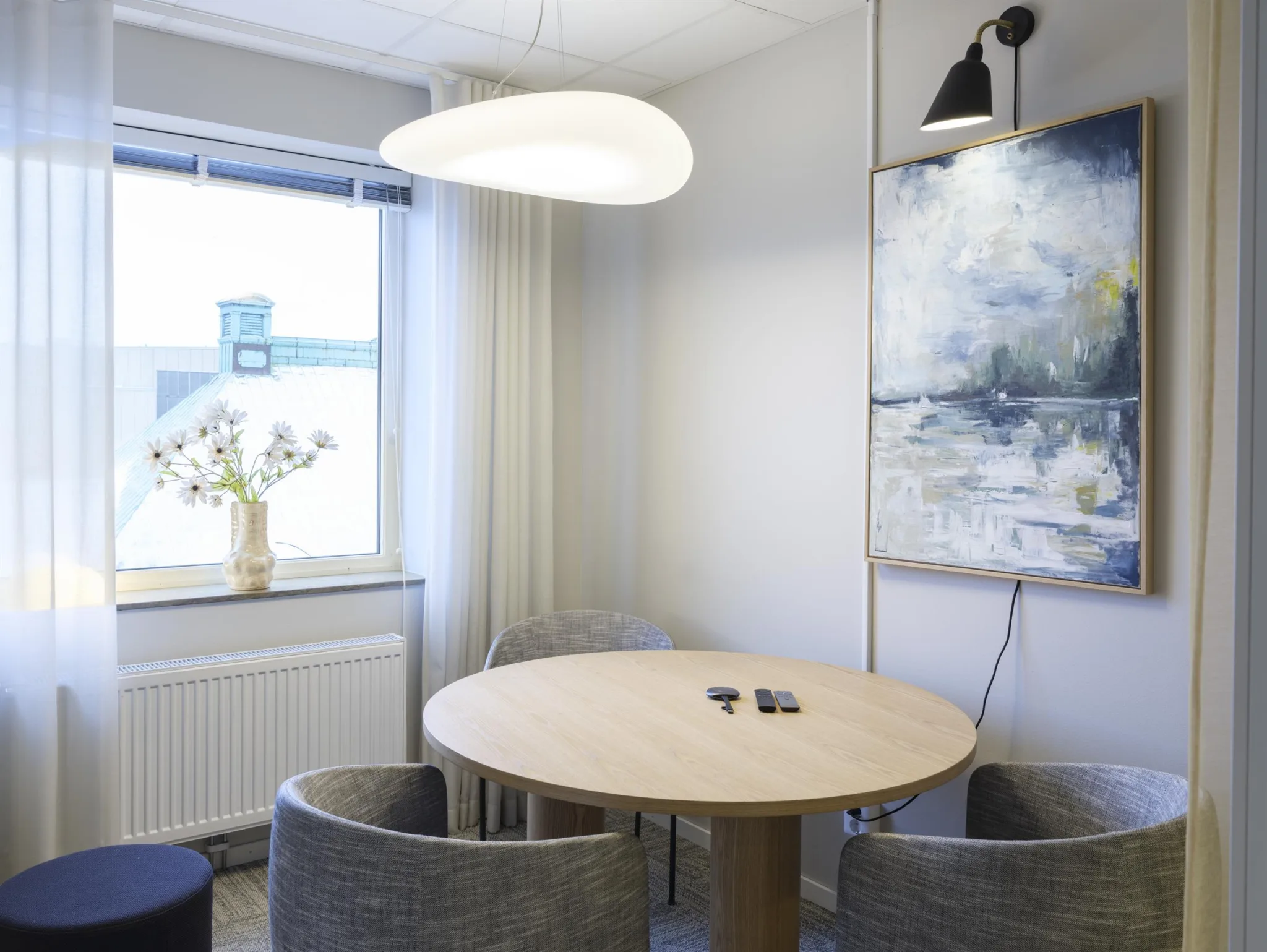 4. Ett litet mötesrum med ett runt träbord, gråa stolar, en abstrakt tavla och ett fönster.