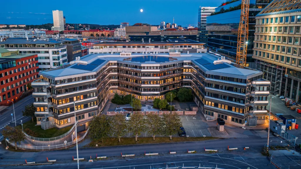 En byggnad med modern arkitektur, omgiven av grönska, stadsmiljö och gatubelysning i skymningen.