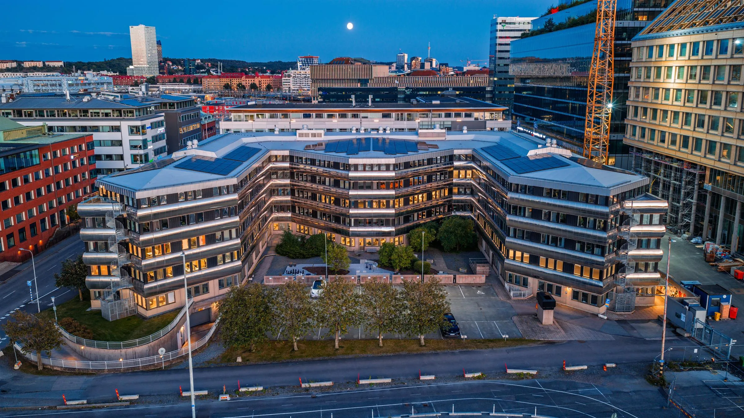 En byggnad med modern arkitektur, omgiven av grönska, stadsmiljö och gatubelysning i skymningen.