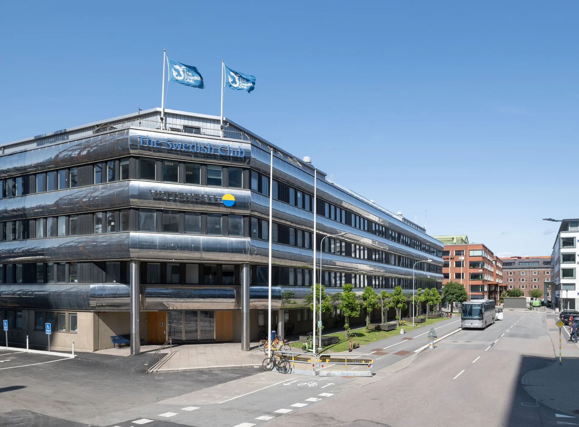 1. En modern byggnad med flera våningar, svensk flagga och två blå flaggor på taket, under en klarblå himmel.