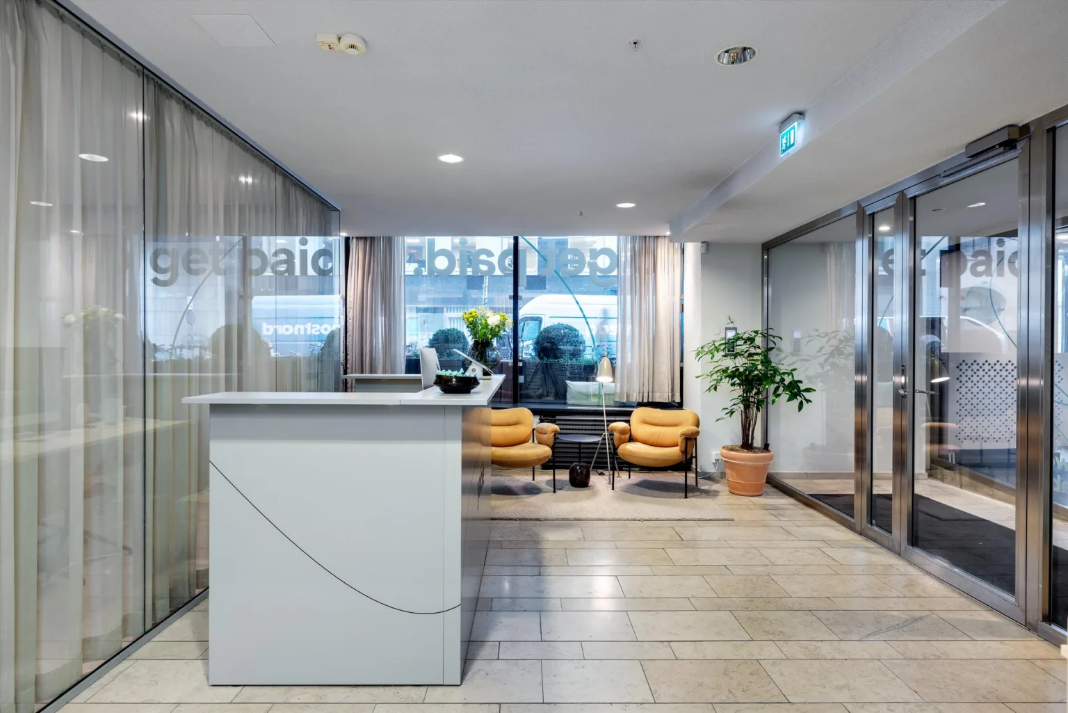 En reception med en vit disk, två gula fåtöljer, växter och glasväggar som vetter mot ett fönster.